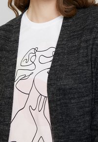 Mörkgrå kofta i mjukt material, öppen fram, som visar en vit t-shirt med en minimalistisk linjeteckning i rosa och svart.