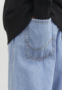 Personne portant un jean en denim bleu clair avec une poche à l'arrière et un haut noir à manches longues, main partiellement dans la poche.