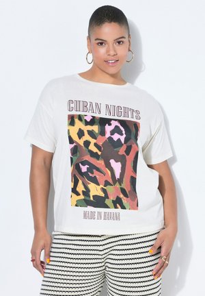 Kobieta z krótkimi włosami, nosząca złote kolczyki w kształcie koła, białą koszulkę z kolorowym, abstrakcyjnym nadrukiem i napisem oraz czarno-białe spodnie w paski.