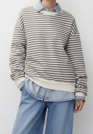 Personne portant un pull rayé gris et blanc par-dessus une chemise en denim clair et un jean bleu clair ample sur un fond uni.