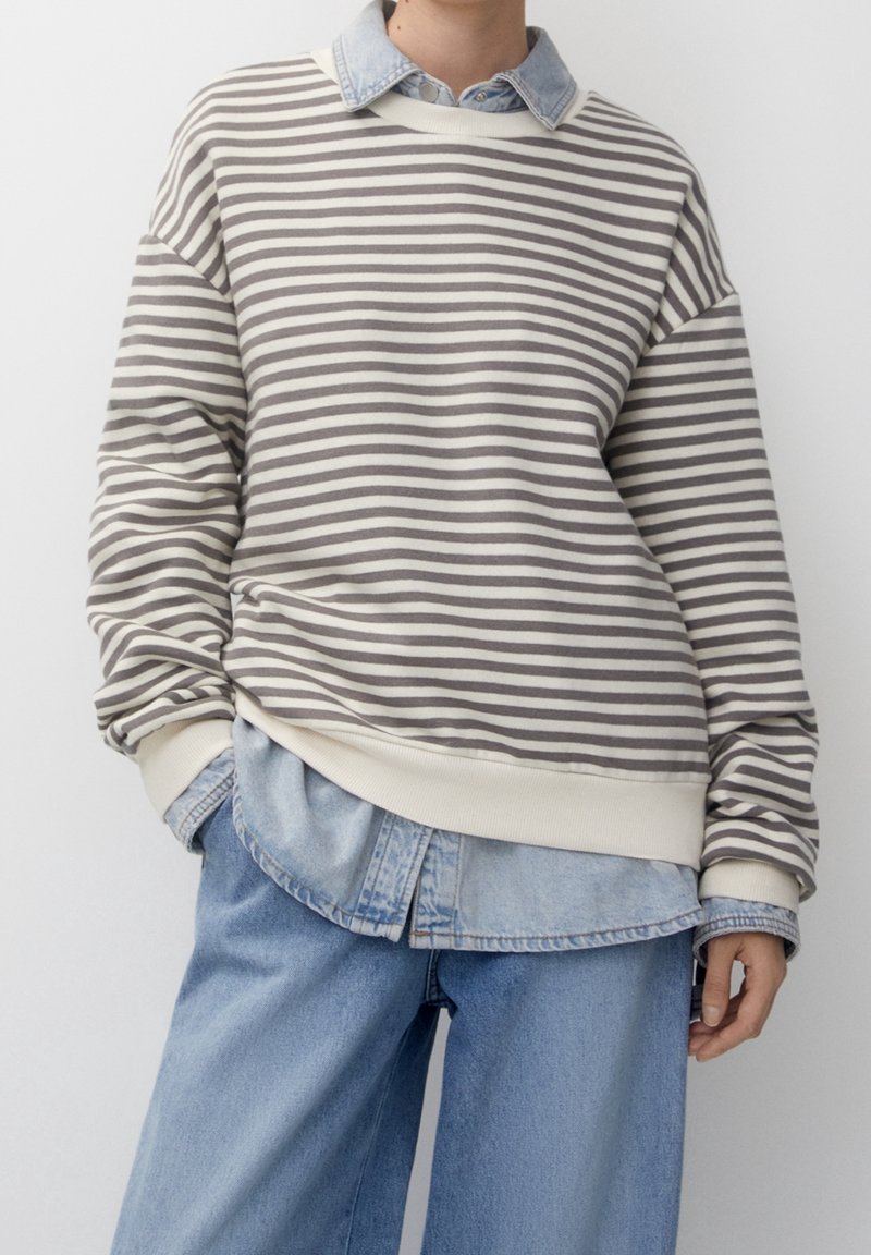 Personne portant un pull rayé gris et blanc par-dessus une chemise en denim clair et un jean bleu clair ample sur un fond uni.