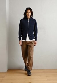 Maglione blu zip con spacchi laterali, camicia bianca sotto, abbinato a pantaloni larghi marroni e scarpe nere su un pavimento di legno.