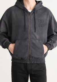 Kapuzensweatjacke in Dunkelgrau, aus weichem Material gefertigt, mit einem gestickten "HUGO"-Logo, elastischen Bündchen und Kängurutaschen.