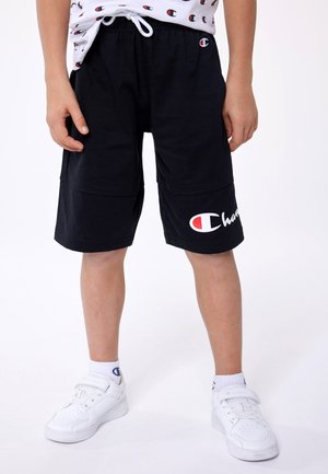 Shorts neri in cotone con tasche laterali, caratterizzati da un logo Champion bianco e rosso sulla gamba sinistra. Abbinati a sneaker bianche e calze.