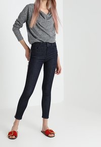 Top lungo a maniche lunghe con collo a cofano grigio; jeans skinny blu scuro; sandali scivolati rossi con un accento dorato. Tessuto texturizzato, design aderente.