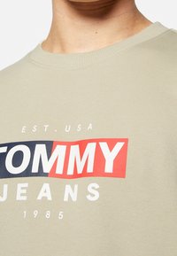 Maglietta beige con scollo rotondo con la scritta "TOMMY" in rosso e "JEANS" in bianco su una striscia blu, tessuto di cotone strutturato.