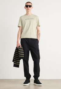 Lichtgroene katoenen T-shirt met "Calvin Klein" graphic, zwarte cargobroek, gestreepte zwart en olijfgroene trui, en zwarte sneakers.