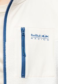 Λευκό μπουφάν με μπλε φερμουάρ και τσέπη, με το λογότυπο "Red Bull Racing" στο στήθος.