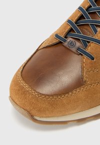 Braune Wildlederschuhe mit einer glatten Lederkappe, marineblauen Schnürsenkeln und einer strukturierten, hellbraunen Sohle. Verfügen über eine Stichnaht entlang der Kanten.
