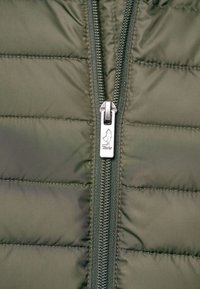 Detailní záběr na zip zelené prošívané bundy se stříbrným jezdcem, který má malý figure logo a nápis "TNCG".