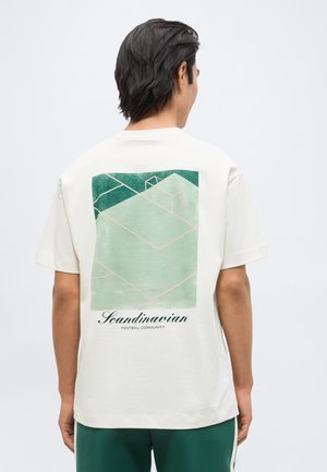 Tume juustega mees, kes kannab valget T-särki, millel on roheline geomeetriline jalgpalliväljakut kujutav muster ja tagaküljel tekst "Skandinaavia Jalgpallikogukond".