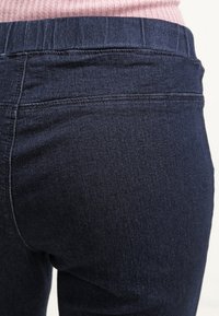 Primer plano de una persona que lleva vaqueros ajustados de mezclilla azul oscuro y una blusa de punto rosa, mostrando la parte trasera de la cintura y la parte superior del muslo.