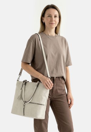 Naine, kes kannab tuhmi hallikat t-särki ja pruune pükse, hoiab suure beeži õlakotiga õlakotiga ja tutidetailiga tavalise valge tausta ees.