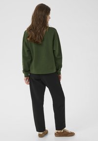 Felpa verde con vestibilità rilassata, polsini e orlo a coste; abbinata a pantaloni neri larghi e sneakers in suede marroni. Design semplice.