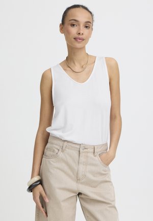 Frau trägt ein weißes ärmelloses Oberteil, beige High-Waist-Hosen, eine goldene Halskette und hält schwarze und weiße Armreifen.