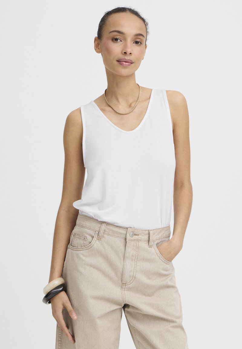 Femme portant un haut blanc sans manches, un pantalon beige taille haute, un collier en or, et tenant des bracelets noirs et blancs.