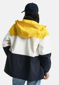 Giacca colorata a tre tonalità in giallo, bianco e blu navy; presenta un cappuccio, una vestibilità ampia e materiale morbido e resistente all'acqua.