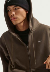 Joven con una sudadera oscura de cremallera y capucha puesta, y un gorro negro de Nike, mirando hacia un lado contra un fondo liso.