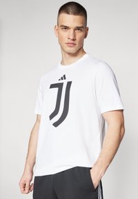 Hvid bomulds T-shirt med et stort sort logodesign. Korte ærmer, rund halsudskæring. Båret med mørke bukser. Enkel og moderne design.