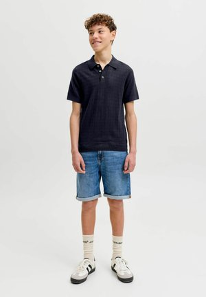 Teenager-Junge steht, trägt ein schwarzes kurzärmeliges Polohemd, hochgekrempelte Jeansshorts, weiße Socken und weiße Turnschuhe mit schwarzen Streifen.