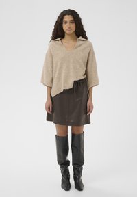Pull beige en mélange de laine avec col en V et col large, associé à une mini-jupe lisse marron foncé et des bottes en cuir noires montant jusqu'au genou.