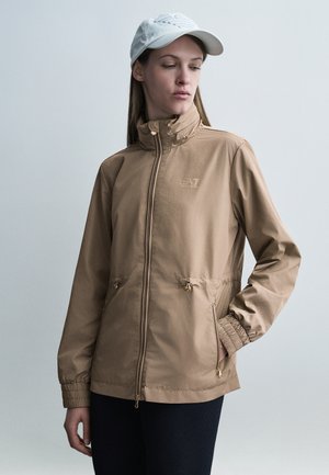 Femme portant une veste beige à fermeture éclair avec des poignets élastiques et une casquette blanche, debout devant un fond clair uni, regardant vers le bas et à gauche.