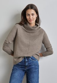 Hellbraunes Cowl-Neck-Pullover aus weichem Material, mit gerippten Bündchen, geradem Schnitt und einem dezenten Nahtdetail auf der Vorderseite. Kombiniert mit blauen Jeans.