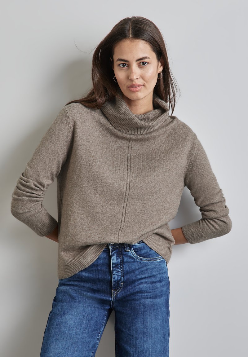 Maglione color marrone chiaro con collo a cowl realizzato in un materiale morbido, caratterizzato da polsini a costine, una vestibilità dritta e un sottile dettaglio di cucitura sul davanti. Abbinato a jeans blu.
