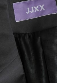 Crni materijal s glatkom teksturom; vidljiva etiketa u ljubičastoj boji s bijelim tekstom "JJXX." Dizajn s nabranim detaljem s jedne strane.