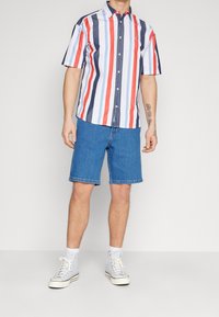 Košile s krátkým rukávem s vertikálními červenými, námořnickými a bílými pruhy; s límečkem na knoflíky. Spárováno s modrými džínovými šortkami po kolena a světlými teniskami.