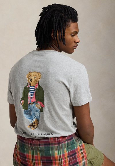 Young man with dreadlocks wearing a gray T-shirt featuring a bear in casual layered clothing printed on the back.
