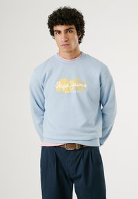 Lichtblauwe sweater met het gele en witte "Pepe Jeans London" logo. Gedragen over een roze shirt, gecombineerd met donkerblauwe broeken en een bruine riem.