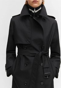 Czarny płaszcz trencz z dwurzędowym zapięciem, dużymi guzikami i paskiem w talii. Charakteryzuje się szerokim kołnierzem i klasycznym krojem.