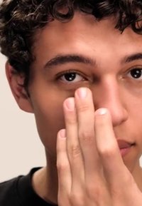 Acerca de un primer plano del rostro de una persona con cabello rizado, aplicando un producto en la zona de debajo de los ojos con los dedos. Fondo neutral, tono de piel natural.