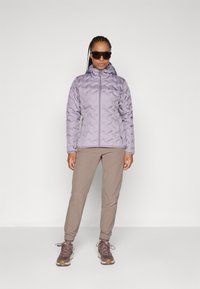 Veste lavande rembourrée avec capuche, fermeture éclair et coutures diagonales, associée à un pantalon beige et des chaussures de randonnée marron. Lunettes de soleil incluses.
