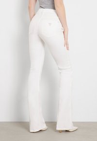 Woman wearing high-waisted white flared jeans and beige heels, standing sideways against a plain light gray wall.
