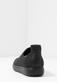 Černá slip-on bota s hladkou texturou, zakřiveným designem a jemnými detaily prošívání. Má polstrovanou podrážku a minimální branding.