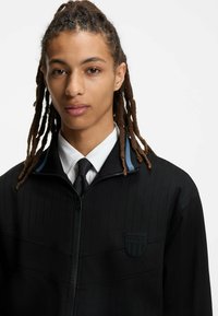 Sort lynlåsjakke med vertikale nålestriber og blå accent på kraven, med tekstureret stof og et HUGO-logo-patch.