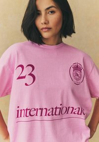 Rozā kokvilnas T-krekls ar numuru "23" tumši burgundijas krāsā un vārdu "starptautisks" tādā pašā krāsā, ar kronīša logotipa detaļu.