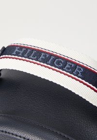 Tume nahktask, millel on valge rihm punaste ja siniste triipudega, embosseeritud "HILFIGER" tekst ja tekstureeritud viimistlus.