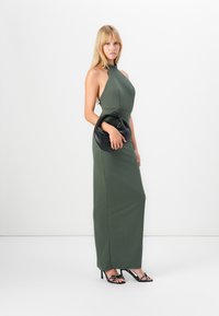 Green halter-neck maxi dress with a fitted silhouette. Paired with a black handbag and black sandals featuring decorative straps.