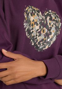 Sudadera morada con un diseño gráfico de corazón que presenta un estampado de leopardos en beige, gris y verde. Textura suave y puños acanalados.