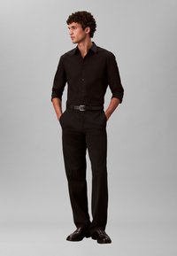 Čierna košeľa na gombíky s dlhým rukávom v kombinácii s čiernymi nohavicami. Oblečenie má elegantný dizajn, priliehavý strih a lesklé čierne topánky.