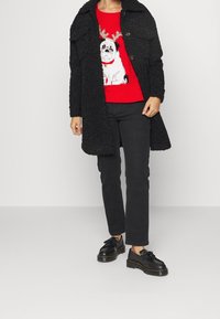 Manteau noir texturé sur un pull rouge avec un chien aux bois de cerf. Jean noir et chaussures noires à semelle épaisse complètent la tenue.