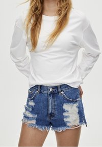 Camisa blanca de manga larga combinada con pantalones cortos de mezclilla azul desgastados con dobladillo desgastado, que presentan desgarros visibles y herrajes metálicos en los bolsillos.