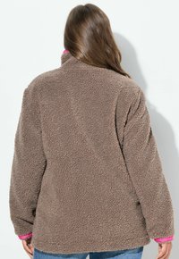 Veste en polaire brune et moelleuse avec un col montant, présentant des accents roses au niveau du col et des poignets. Coupe large et surface texturée.