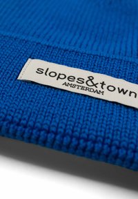 Berretto di maglia blu con un motivo testurizzato. Presenta un'etichetta in tessuto color crema con la scritta "slopes&town AMSTERDAM" in caratteri neri.