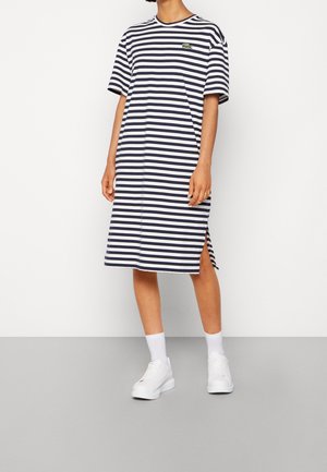 Persoon draagt een knielange, donkerblauw-wit gestreepte jurk met korte mouwen en een zijspalk, witte sokken en witte sneakers, staand op een grijze vloer.