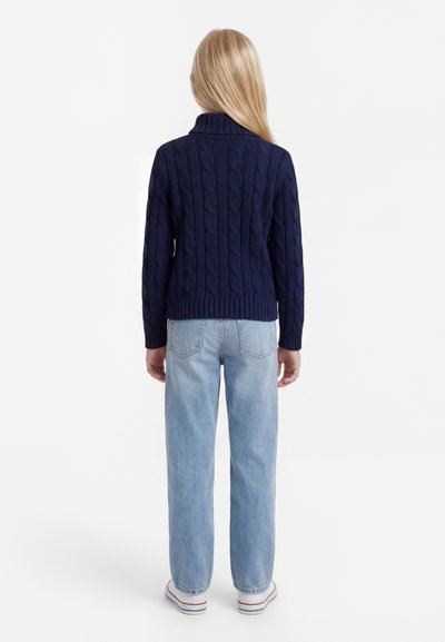 Otrok z dolgimi blond lasmi, ki gleda proč, oblečen v mornarsko modro pleten pulover, svetlo modre kavbojke in bele superge na belem ozadju.