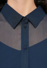 Μπλε μπλούζα με διάφανο πάνελ και στερεό γιακά. Διαθέτει πατιλέτα με στρογγυλά κουμπιά. Διακριτή υφή εμφανής στο ύφασμα.